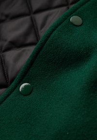 Tejido de mezcla de lana verde con un acabado texturizado, combinado con un forro negro acolchado; cuenta con botones verde brillante en el borde.
