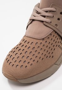 Zapato deportivo marrón con parte superior de gamuza texturizada que presenta patrones perforados, cuello de tela y cordones planos, todo en un diseño monocromático.