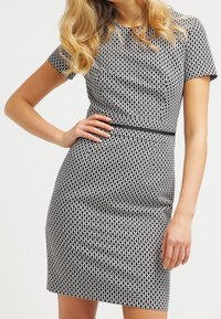 Robe à motif noir et blanc, silhouette ajustée, manches courtes et taille marquée. Tissu lisse, détails de design géométrique.