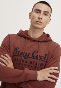 Felpa a cappuccio color bordeaux con cordini, con testo stampato in nero. Il tessuto appare morbido, con una vestibilità comoda e polsini a costine.