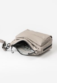 Lichtgrijze crossbody tas met open rits die een binnenzak onthult, en een verstelbare schouderband met zwarte clips op een witte achtergrond.