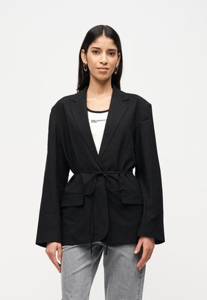Femme portant un blazer noir ceinturé, un haut blanc avec du texte, un jean gris clair et des boucles d'oreilles créoles dorées, sur fond blanc.