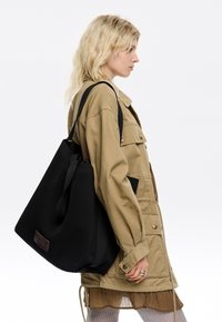 Zwarte mesh schoudertas met schouderbanden, met een gestructureerde afwerking en een bruine leren logo-patch. Draag ik door iemand in een beige jas.