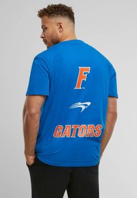 Sinine puuvillane T-särk, millel on oranž "F" logo ja "GATORS" tekst seljal, tekstist allpool valge graafika. Lühikesed varrukad.