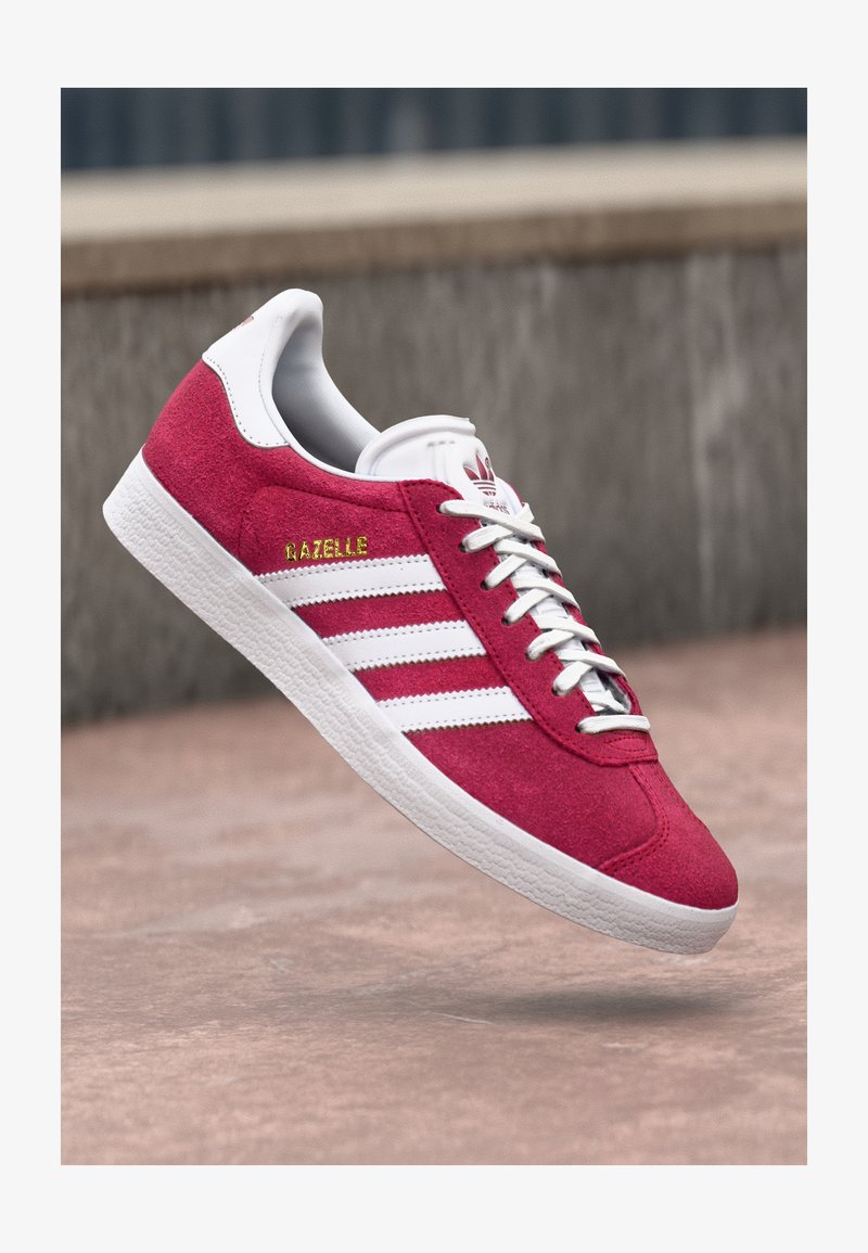 Rdeče semiš športne čevlje z belimi usnjenimi črtami. Imajo zlato oznako "Gazelle" in teksturiran gumijast podplat. Klasična zasnova.