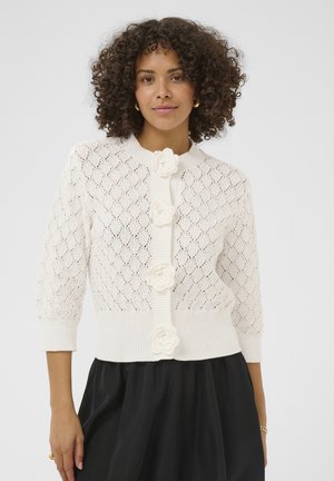 Kvinde med krøllet hår iført en hvid strikcardigan med blomsterknapper og et sort nederdel, poserer mod en ensfarvet baggrund.