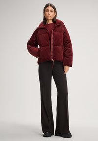 Giacca a vento bordeaux con colletto alto, texture trapuntata, zip frontale e orlo con cordino, abbinata a pantaloni neri a campana e stivali.