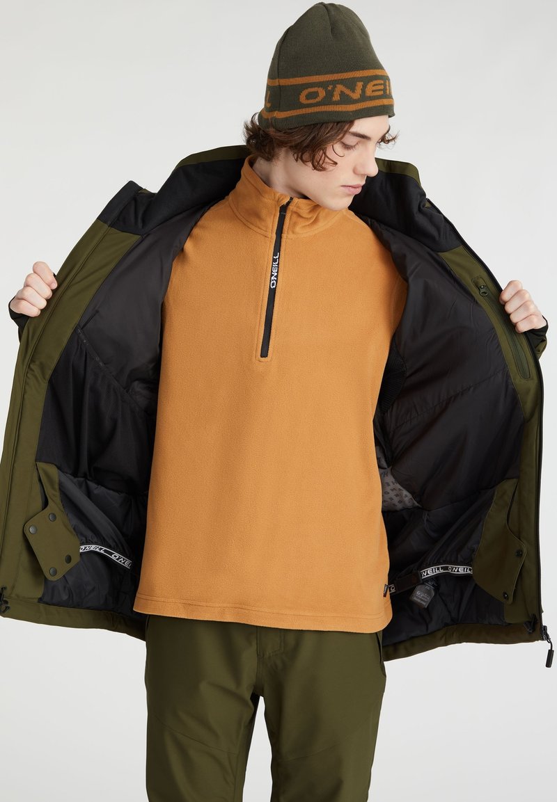 Oranger Fleece-Pullover mit Reißverschlusskragen, getragen unter einer schwarz-grünen Jacke. Das Model trägt zudem olivgrüne Hosen und eine passende Mütze.