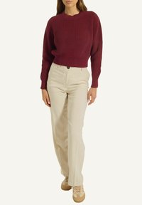 Frau, die einen bordeauxroten Rippstrickpullover, beige hochgeschnittene gerade Hose und beiges Sneakers trägt, steht vor einem einfarbigen Hintergrund.