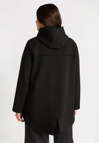 Svart huva kappa med avslappnad passform, med rak skärning, detalj i ryggen och mjukt material. Slät tyg med minimal accent.