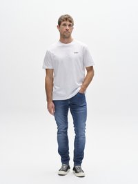 Balti trumpomis rankovėmis marškinėliai su juodu logotipu, derinami su mėlynomis aptempto silueto džinsais ir juodai baltais sportbačiais, stovintys prieš paprastą foną.