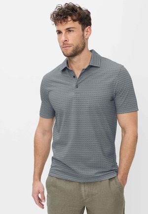 Mann mit lockigem Haar, der ein kurzärmeliges, grau gemustertes Poloshirt und helle olivgrüne Hosen trägt, steht mit einer Hand in der Tasche.