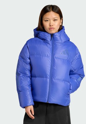 Junge Frau trägt eine glänzende blaue Steppjacke mit Kapuze, steht vor einem hellgrauen Hintergrund.