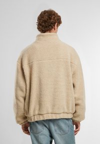 Giacca in pile beige con colletto alto, spalle scese e superficie morbida e testurizzata. Presenta un orlo dritto e una vestibilità rilassata.