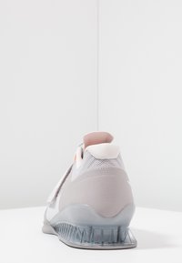 Grå atletisk sko med mesh-overdel, polstret hæl, transparent sål og orange detalje nær bagenden. Solidt design, tekstureret finish.