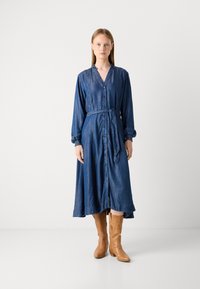 Denim srajčna obleka z dolgimi rokavi, z gumbnim zapiranjem in pasom. Ima razširjeno krilo, temno modro barvo in mehak teksturo. Rjave kavbojske čevlje.