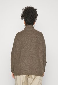 Pull marron tricoté avec un col montant, des épaules tombantes et une coupe ample, fabriqué en tissu texturé. Coutures visibles sur les côtés.