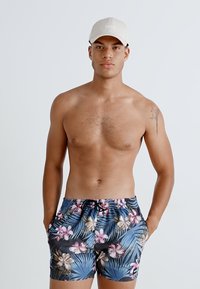 Bărbat fără cămașă, purtând o pălărie bej și șorturi de baie cu model floral, cu mâinile în buzunare, stând în fața unui fundal simplu.