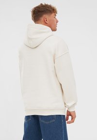 Sudadera beige claro hecha de una tela suave, con una gran capucha, corte holgado y puños acanalados. Combinada con jeans azules.