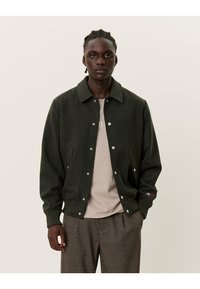 Giacca in misto lana verde scuro con chiusura a bottoni, colletto oversize e tasche laterali. Indossata sopra una camicia beige e pantaloni a fantasia.