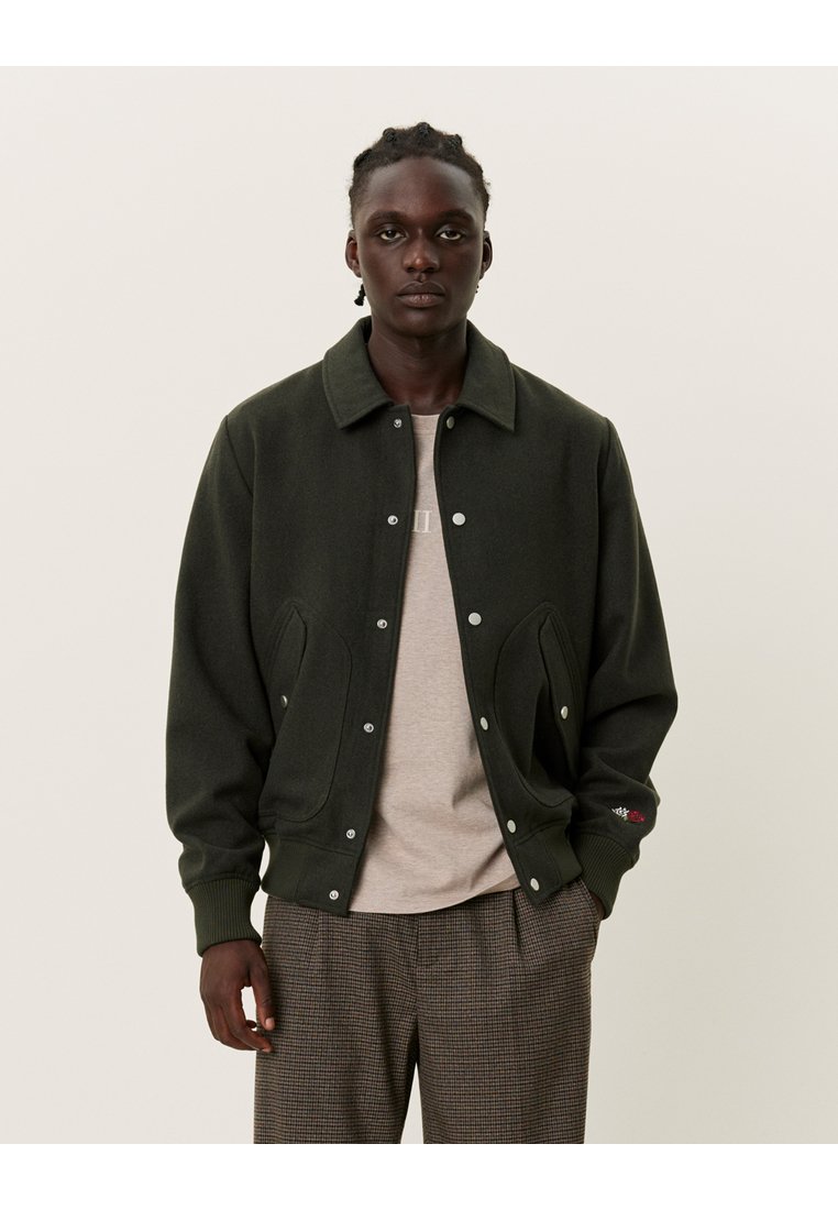 Giacca in misto lana verde scuro con chiusura a bottoni, colletto oversize e tasche laterali. Indossata sopra una camicia beige e pantaloni a fantasia.