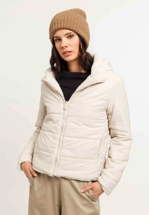 Chaqueta acolchada beige clara con capucha y forro de piel sintética, con cierre de cremallera en la parte frontal y bolsillos laterales, acompañada de un gorro de lana marrón.