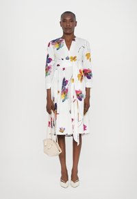 Kukkakuosiset mekko valkoisella taustalla, V-pääntie, kolme neljäsosaa hihat, kireä vyötärö ja liukuva hame. Beige laukku ja korot.