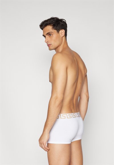 White boxer briefs made from smooth fabric, featuring a gold Greek key design on the waistband. The fit is snug and contoured.