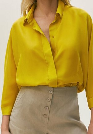 Kobieta ubrana w luźną, jasnżółtą koszulę na guziki, wpuszczoną w wysokowojne beżowe spodnie z czterema guzikami z przodu.