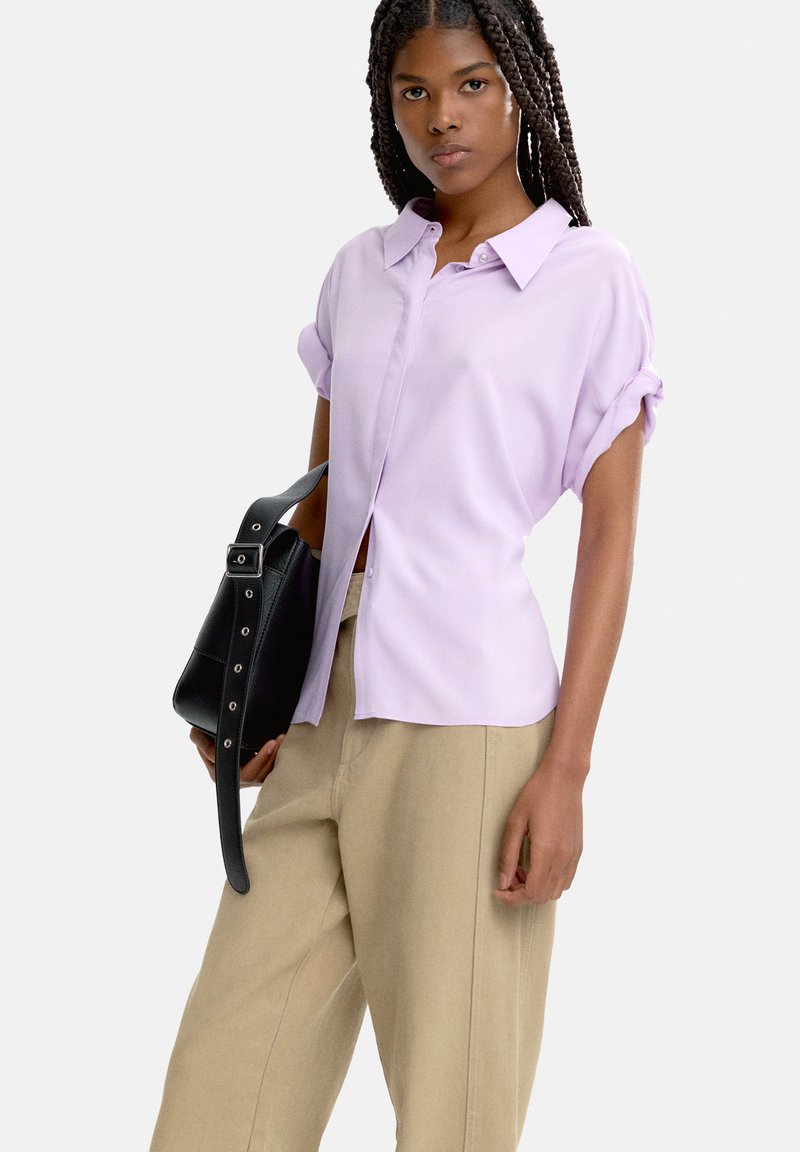 Jasnofioletowa koszula z krótkim rękawem i zapięciem na guziki, zestawiona z beżowymi spodniami o wysokim stanie; nosząca czarną skórzaną torbę na ramię.