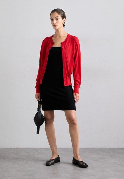 Cardigan rosso, abito aderente nero, ballerine nere intrecciate e una borsa rotonda nera. Texture lisce e una silhouette classica. Chiusura a bottoni semplice.