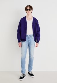 Lilla strikket cardigan med svarte knapper og striper, lagt over en hvit tank topp. Lyseblå jeans og svarte joggesko fullfører antrekket.