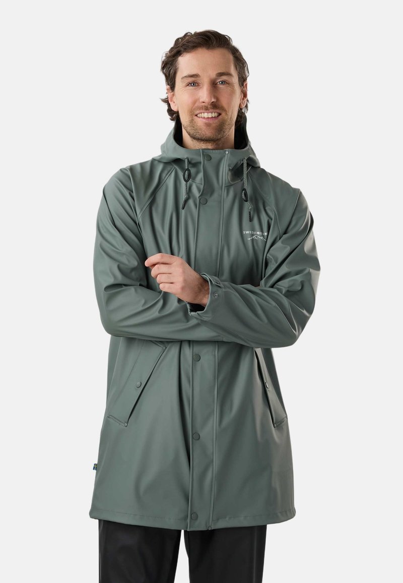 Uomo sorridente che indossa una giacca impermeabile verde con cappuccio, tasche frontali e coulisse regolabili, in piedi contro uno sfondo uniforme.