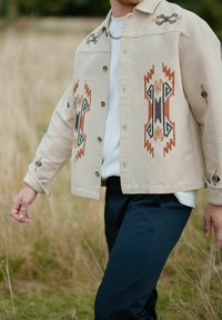 Beige jas met geborduurde geometrische patronen in bruin, zwart en teal; heeft knopen en een kraag. Gedragen over een wit shirt en donkere broek.