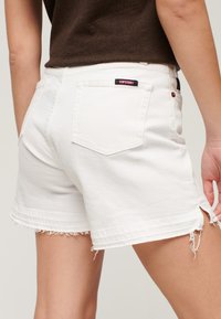 Shorts de mezclilla blanca con dobladillos deshilachados, dos bolsillos traseros y una etiqueta de "Superdry" en negro y rojo en la cintura. Diseño de corte recto.