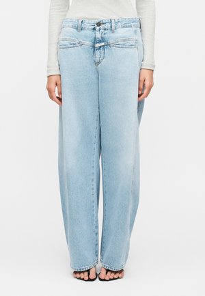 Osoba nosząca jasnoniebieskie szerokie jeansy z podwójnym paskiem w talii, w połączeniu z czarnymi sandałami na obcasie z odkrytymi palcami i lekką bluzką z długim rękawem.