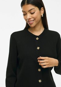 Cardigan en tricot noir avec un col rond, comportant six boutons ronds de couleur claire et des manches longues. Texture lisse et design ajusté.