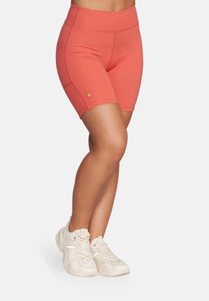 Kobieta w koralowych, wysokich spodenkach rowerowych i beżowych tenisówkach, z nogami skrzyżowanymi na białym, jednolitym tle.