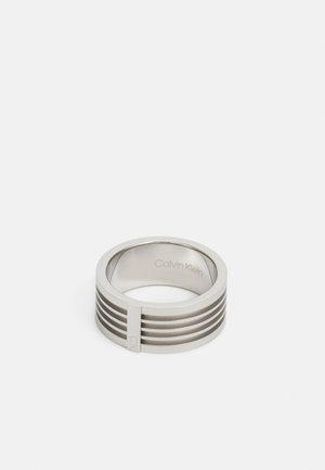 Stainless steel ring featuring a smooth finish with horizontal grooves and a small integrated accent. Engraved brand name inside.