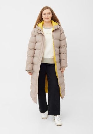 Lange gewatteerde jas in lichtbeige met een gele voering, voorzien van een capuchon, ribgebreide boorden en voorzakken. Gedragen over een licht sweatshirt en zwarte broek.