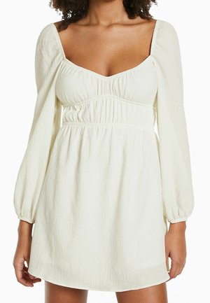 Femme portant une mini-robe blanche texturée avec des manches longues bouffantes et un décolleté carré.