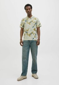Camisa polo en verde pálido con un patrón de hojas en amarillo, azul y gris. Manga corta, cuello, combinada con jeans azul claro y zapatos beige.
