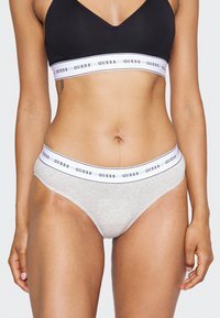 Soutien-gorge noir avec un design en V associé à un sous-vêtement gris featuring une bande élastique blanche avec "GUESS" imprimé en bleu marine. Matière en coton doux.