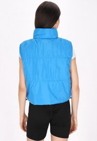 Vest fără mâneci, albastru, tip puffer, cu guler înalt, textură lucioasă, tiv elastic și cusături orizontale, purtat peste o cămașă albă și pantaloni scurți negri.