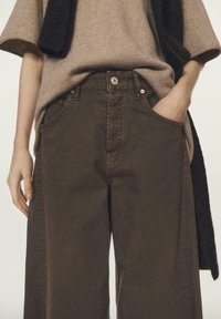 Brązowe spodnie o szerokich nogawkach z teksturowanym wykończeniem, z przednimi kieszeniami, zapięciem na guziki i delikatnym połyskiem. Połączone z beżowym dzianinowym topem.