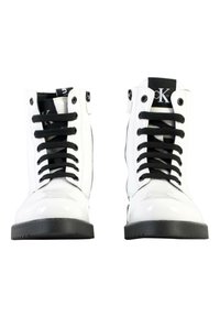 Botas altas blancas con acabado brillante, cordones negros y un acento negro en la lengua que presenta la letra "K". Suelas de goma con agarre texturizado.