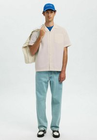 Korte mouwen licht geruit overhemd, blauwe T-shirt eronder, lichtblauwe jeans, zwart-witte schoenen, draagt een beige tas, blauwe pet.