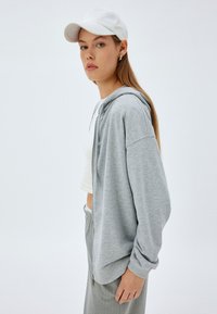 Sudadera con cremallera de color gris claro, con un corte amplio, mangas largas y capucha. Lleva puesta sobre una camiseta corta blanca, combinada con pantalones rayados en gris claro.