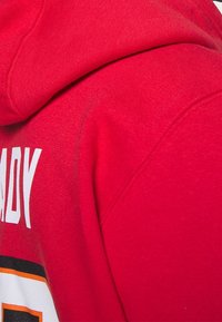 Roter Kapuzenpullover mit weißem Text, elastischem Material und einer Kängurutasche. Der Text beinhaltet einen Namen und ein auffälliges Zahlen-Design.