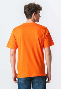 Ung mand med kort, krøllet hår iført en enkel, lys orange kortærmet T-shirt og blå jeans, set fra ryggen mod en hvid baggrund.